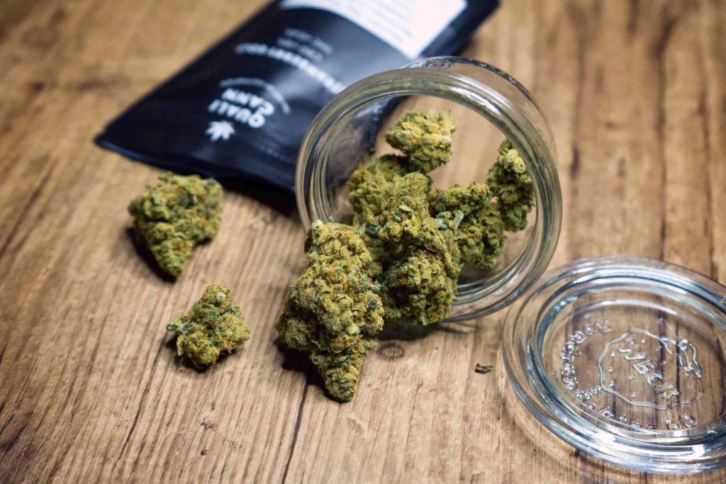 Glas mit hochwertigen CBD-Blüten, das auf einem Holztisch liegt, während einige Blüten herausfallen. Im Hintergrund eine schwarze Verpackung mit Qualicann-Branding