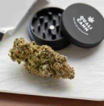 Qualicann CBD-Blüte auf weißem Untergrund, im Hintergrund ein geöffneter schwarzer Grinder mit Logo und eine Packung Papers.