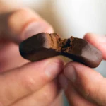 Nahaufnahme zweier Finger, die ein Stück weiches, braunes CBD-Haschisch auseinanderbrechen – feinporige, harzige Textur im Fokus.