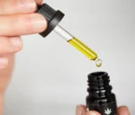 Hand hält eine Pipette mit goldenem CBD-Öl über eine geöffnete schwarze Flasche mit Cannabis-Logo, Tropfen fällt herunter.