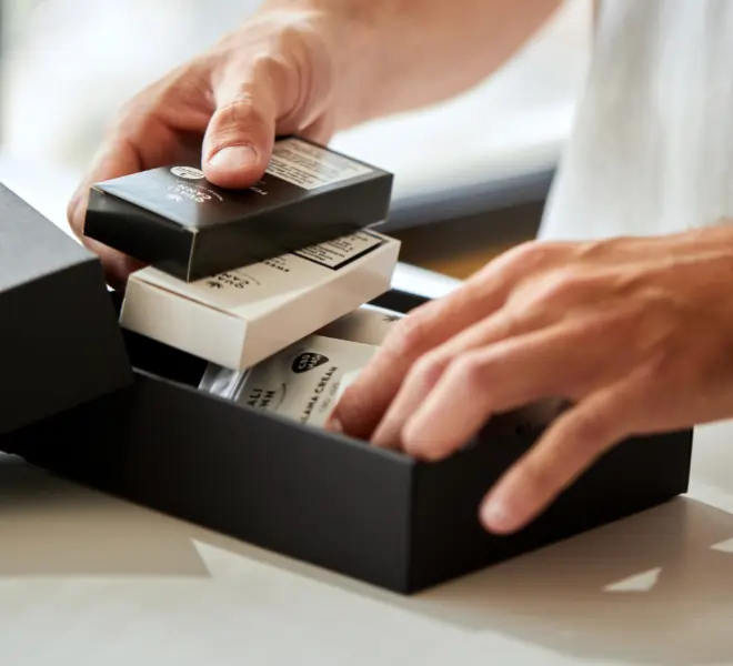 Zwei Hände packen eine schwarze Box mit weißen und schwarzen Produktverpackungen, die sorgfältig hineingelegt werden. Das Bild vermittelt den Eindruck von hochwertiger, stilvoller Verpackung.