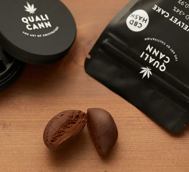 Ein rundliches, aufgebrochenes Stück CBD Hash auf einem Holztisch. Daneben sieht man eine schwarze Verpackung sowie einen Grinder-Deckel mit dem Logo von Qualicann