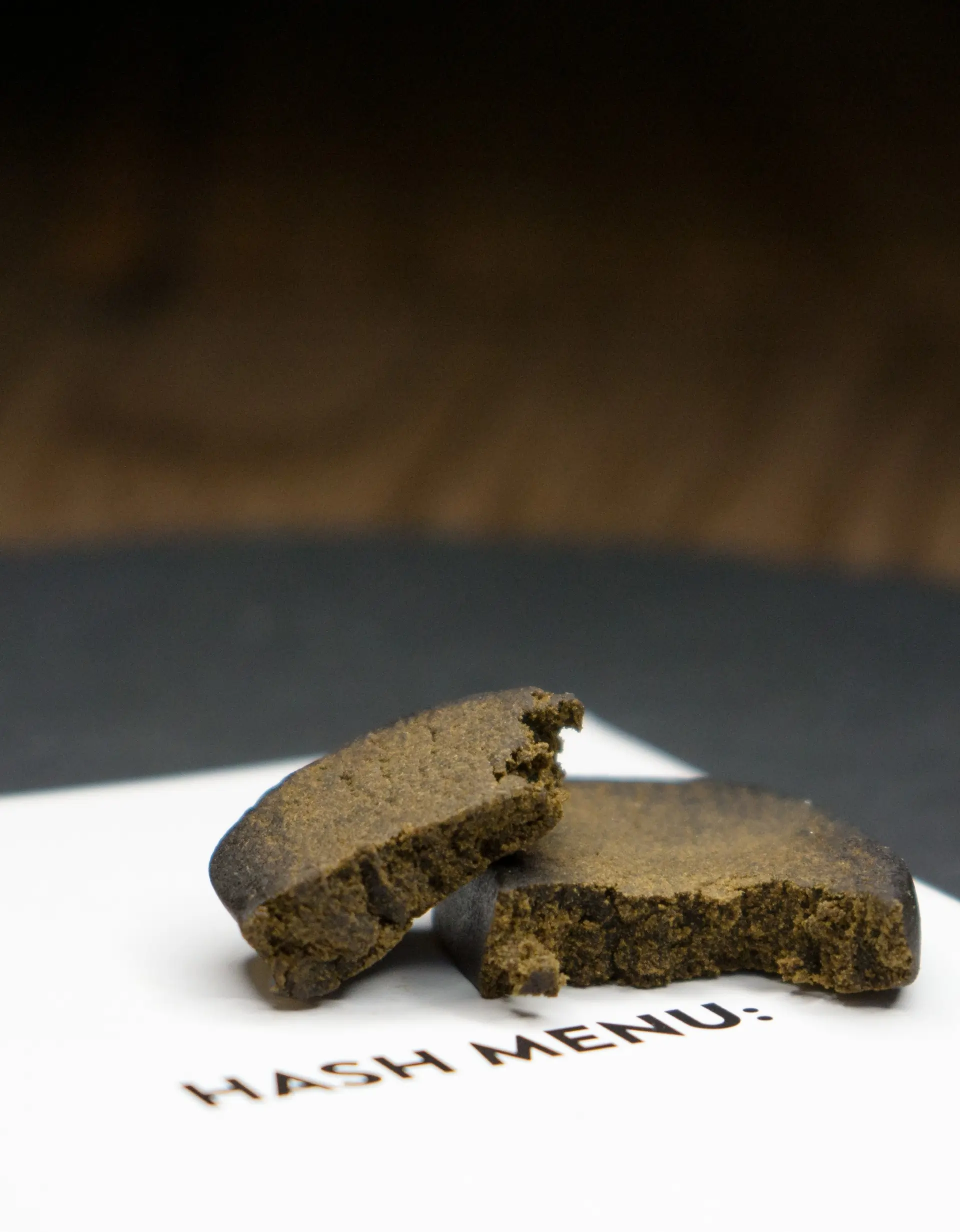 Zwei Stücke von braunem, gepresstem CBD Hash zu sehen, die auf einem weißen Untergrund mit der Aufschrift „HASH MENU:“ liegen. Die Konsistenz wirkt fest, aber bröckelig
