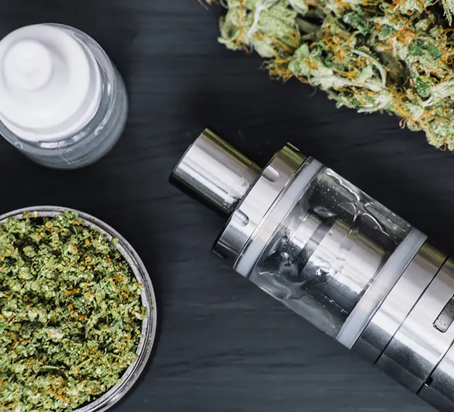 Ein moderner CBD Vaporizer aus Edelstahl, daneben fein zerkleinerte CBD Blüten sowie weiteres Zubehör. Die Szene vermittelt ein hochwertiges und professionelles Setup für das Vaporisieren von CBD.