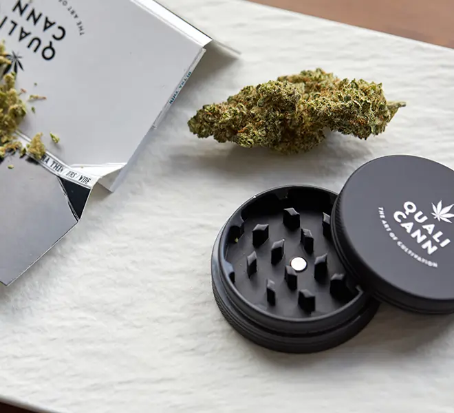 Nahaufnahme eines Qualicann Grinders aus Aluminium neben einer CBD-Blüte und Produktverpackung – steht für hochwertiges Zubehör und Premiumqualität bei CBD-Produkten.