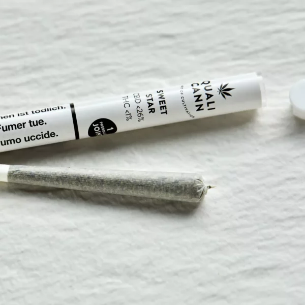 Ein vorgedrehter CBD Joint liegt auf einer weißen Steinplatte, begleitet von seiner Verpackung - einem weißen Plastikröhrchen und einem Gummideckel